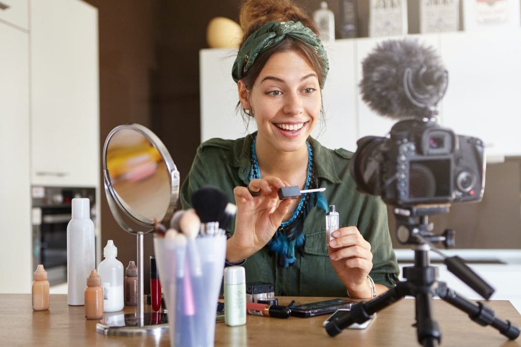 Female video channel host using lip gloss for putting make up, telling about cosmetics to her custom